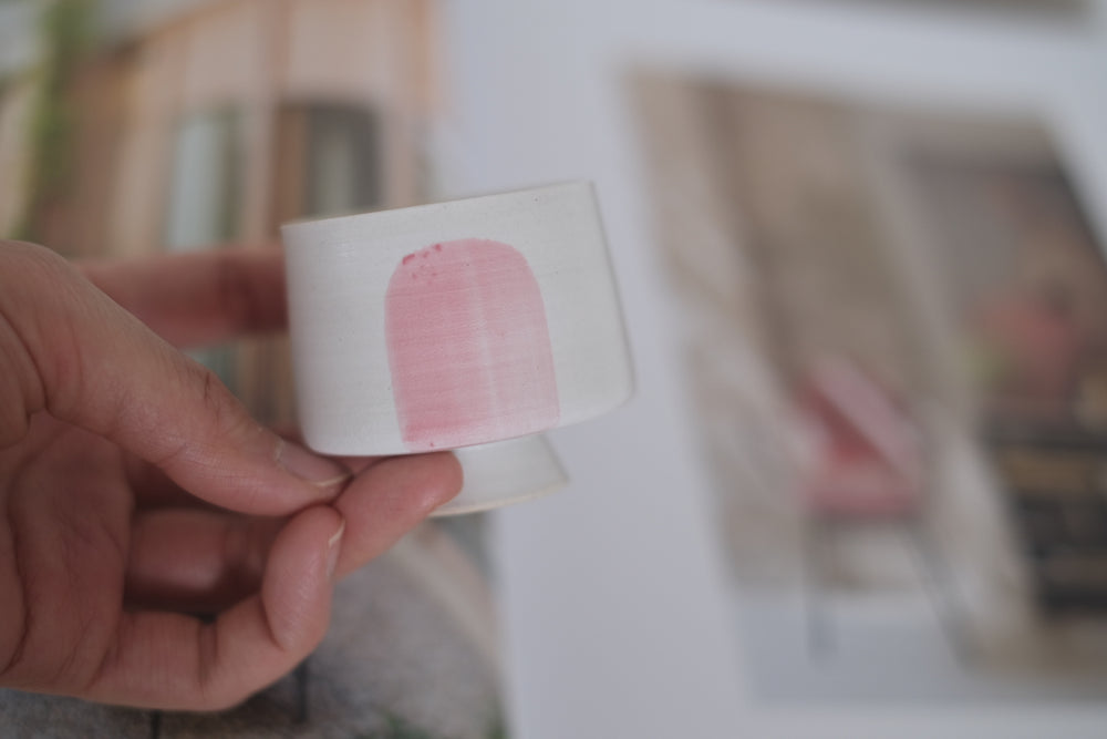 A close-up image shows the Chinese ceramic cup held gently in a hand, highlighting its petite size and delicate pink brushstroke detail. The soft, unglazed texture adds a rustic feel to the minimalist design.