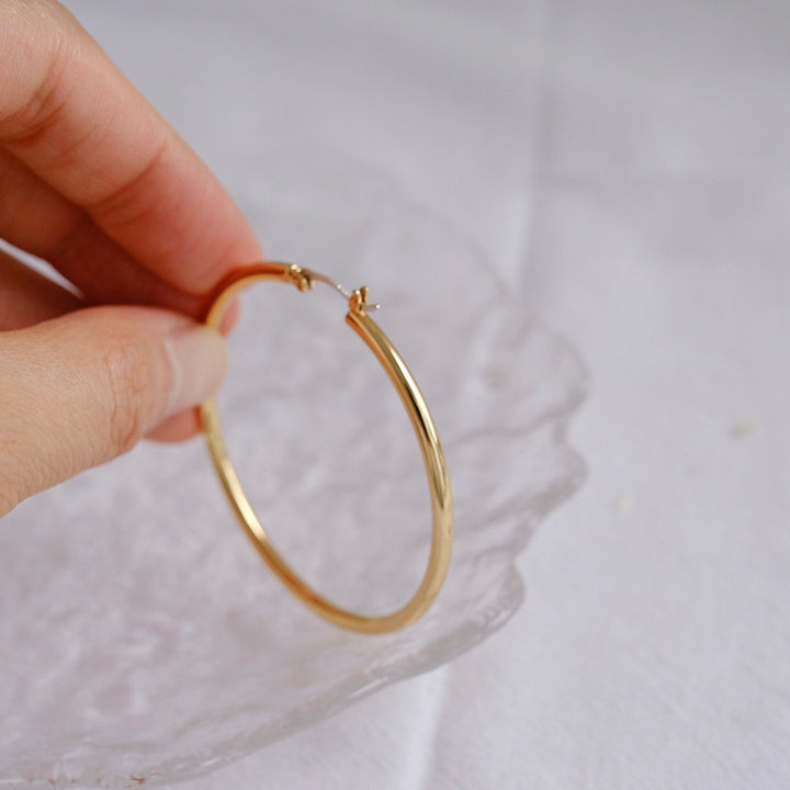 A close-up of a hand holding one hoop earring, highlighting its sleek design, secure clasp, and luxurious golden shine.