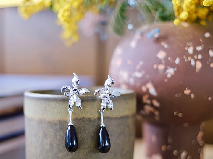 The Daffodil earrings displayed on a ceramic stand with a blurred floral background, highlighting the shiny silver-plated brass and the elegant black teardrop dangle.