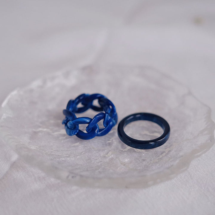 The dark blue and light blue rings are artistically displayed on a clear glass plate. This close-up emphasizes the fine details and craftsmanship of the interwoven ring, as well as the smooth finish of the band, making them a captivating duo.
