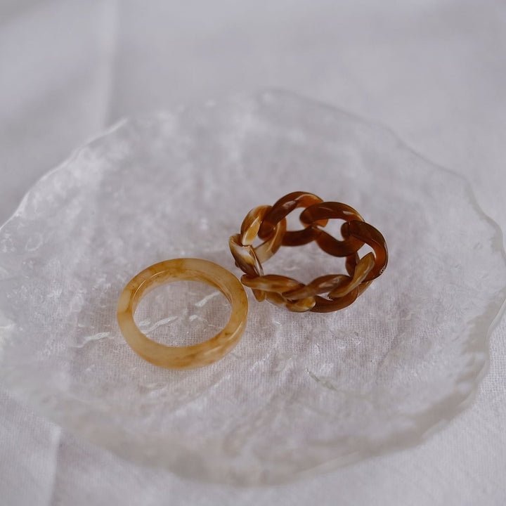 This image highlights the beige and coffee-colored rings placed on a delicate glass plate. The intricate weave of the coffee-colored ring and the sleek surface of the beige band are accentuated by the light, showcasing their refined design and elegant colors.