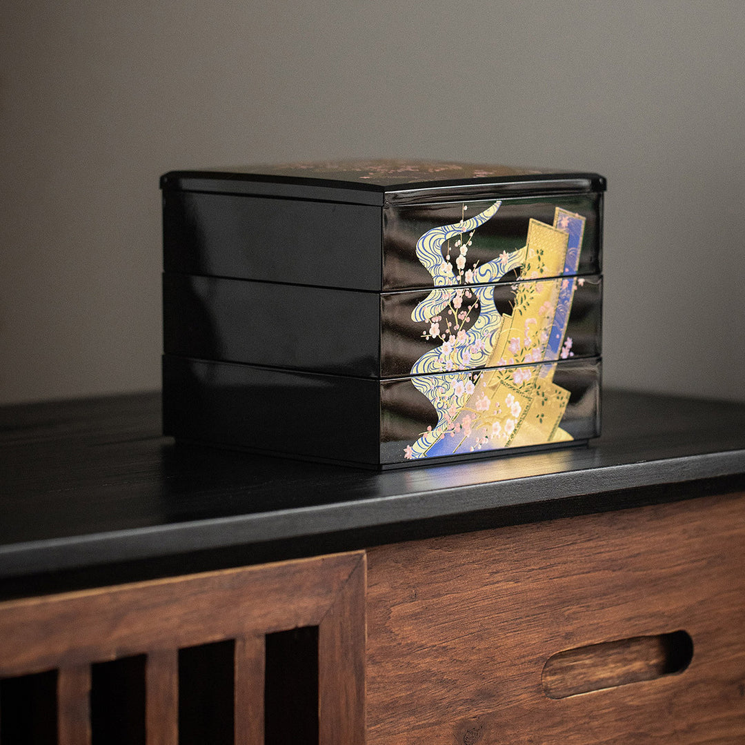 A close-up view of the jewelry box focusing on the delicate gold leaf artwork. The intricate patterns of kimonos and cherry blossoms are displayed in stunning detail, capturing the essence of traditional Japanese artistry. The lustrous lacquer finish enhances the depth and richness of the gold leaf, making the designs stand out against the dark background.