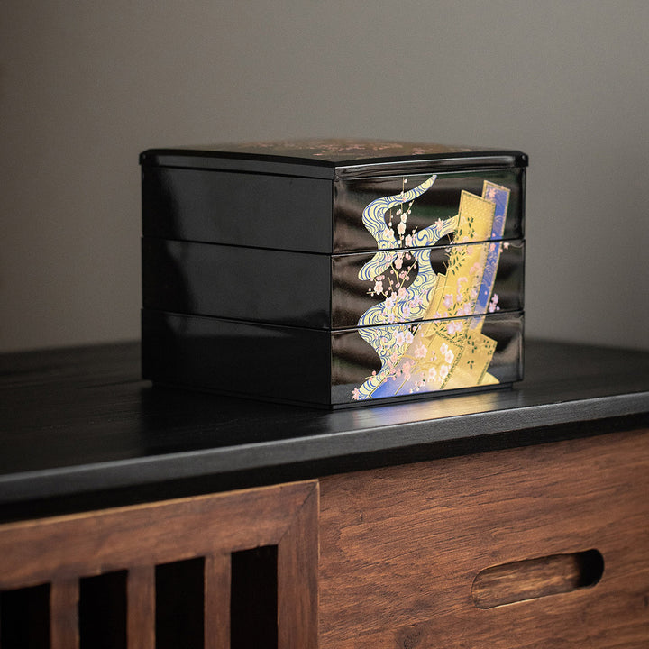 A close-up view of the jewelry box focusing on the delicate gold leaf artwork. The intricate patterns of kimonos and cherry blossoms are displayed in stunning detail, capturing the essence of traditional Japanese artistry. The lustrous lacquer finish enhances the depth and richness of the gold leaf, making the designs stand out against the dark background.