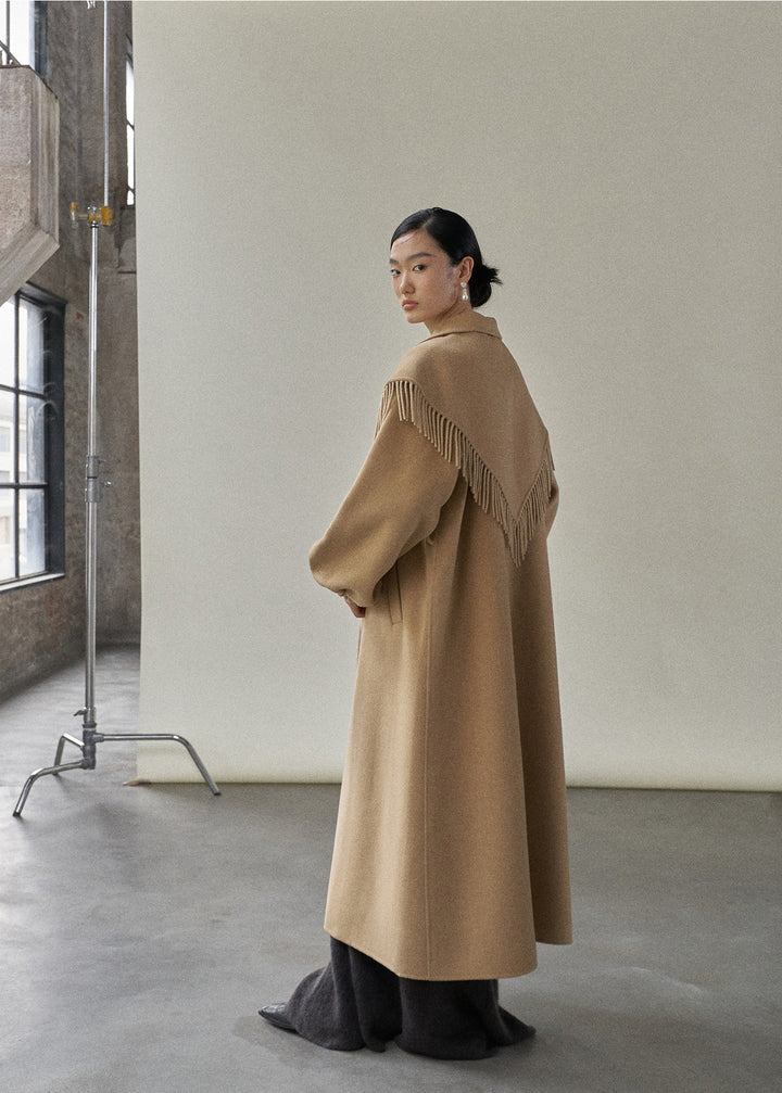 Side profile of the model in the camel coat, demonstrating its long length and sophisticated silhouette.