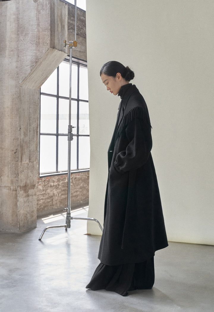 Side profile of the model in the black coat, displaying the coat’s elegant length and structure.