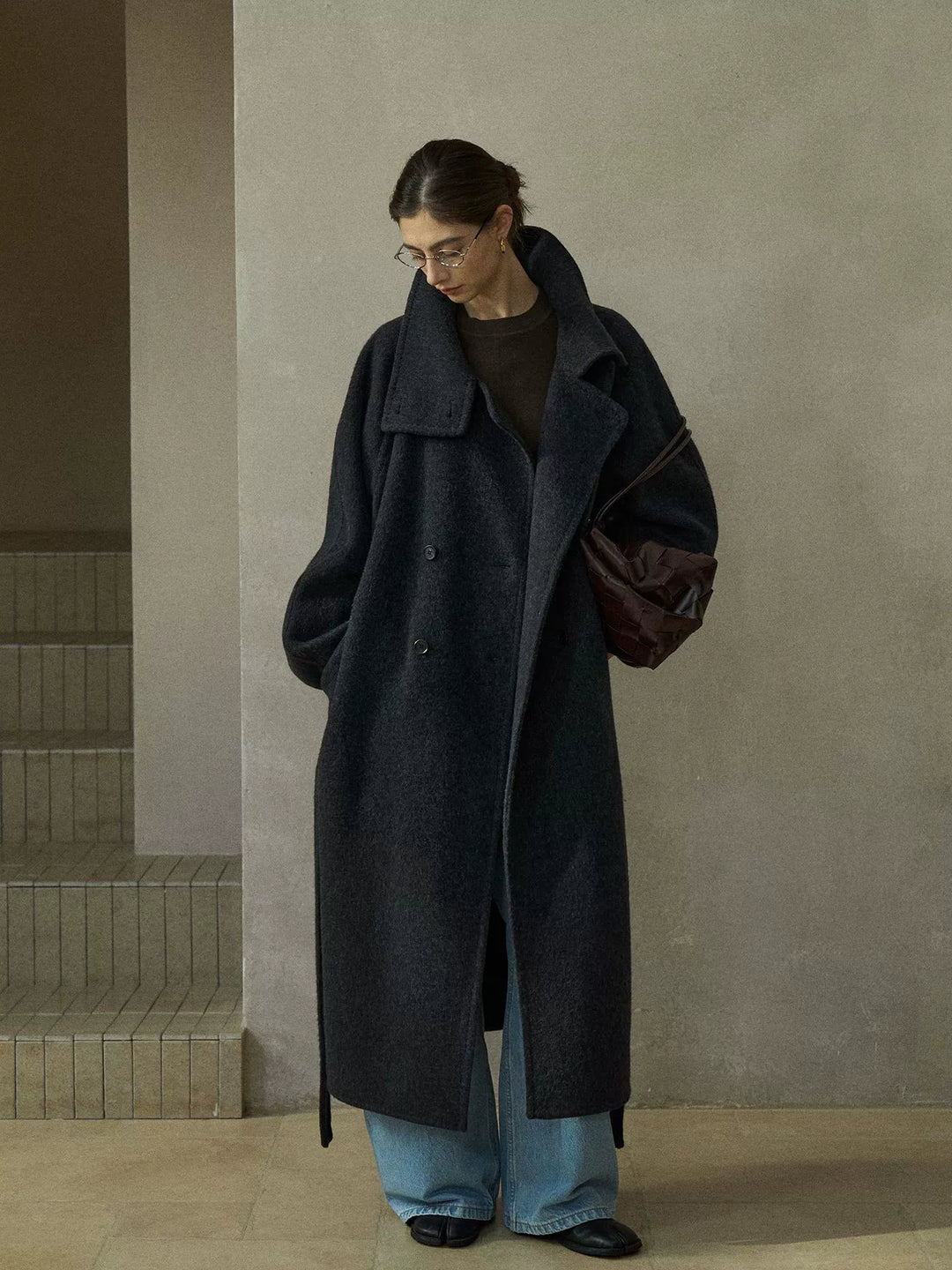 Model wearing charcoal grey wool coat with oversized collar, double-breasted button closure, and tie belt, styled with light blue jeans