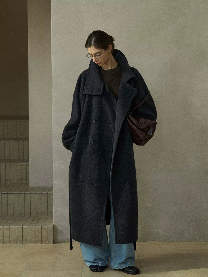 Model wearing charcoal grey wool coat with oversized collar, double-breasted button closure, and tie belt, styled with light blue jeans