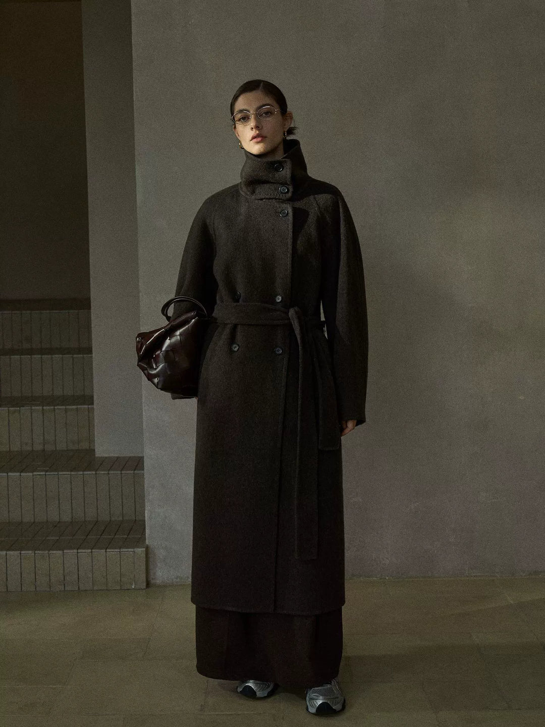 Model wearing chocolate brown wool coat with oversized collar, double-breasted button closure, and tie belt